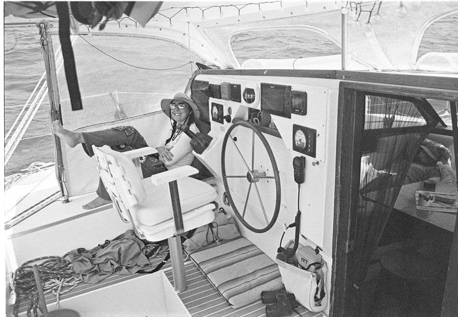 Black and white photo of Sam relaxing in a sun hat at the helm of a sailboat, feet up beside the wheel