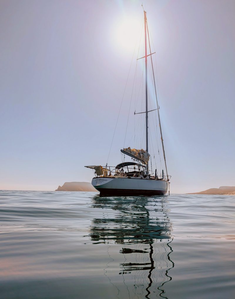 Sailboat anchored on glassy water with its mast reflected below and the sun glowing overhead